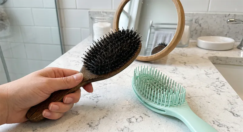 A person brushing their hair in front of a bathroom mirror with a paddle brush visible in the reflection