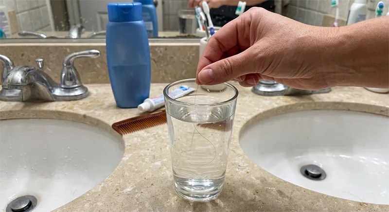 A single hair strand floating in a clear glass of water during a hair porosity float test on a bathroom countertop