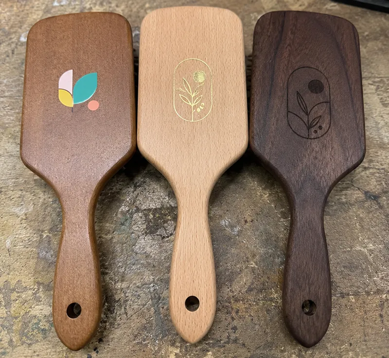 Three paddle brushes showing different logo application methods — pad printing, hot stamping, and laser engraving on handles