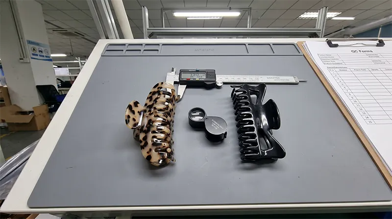 Cellulose acetate hair clip and ABS plastic hair clip placed side by side on a factory inspection desk showing material thickness and colour differences